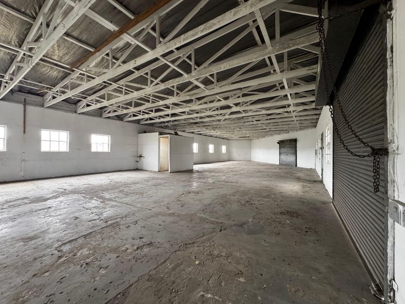 Spacious industrial warehouse area featuring exposed steel roof trusses, concrete flooring, and multiple roller shutter doors, offering a versatile open-plan workspace.