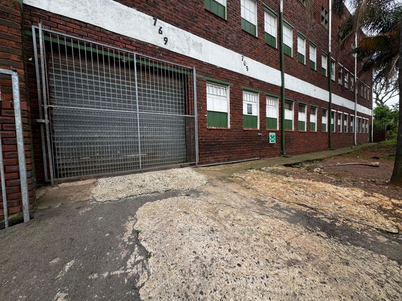 Industrial building exterior featuring a red brick facade, roller shutter access, and multi-pane windows with visible wear to the surrounding concrete driveway.