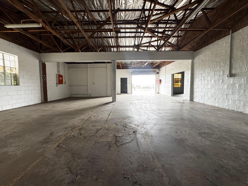 Spacious industrial warehouse unit featuring exposed timber roof trusses, concrete flooring, painted brick walls, and a large roller shutter door for easy access.