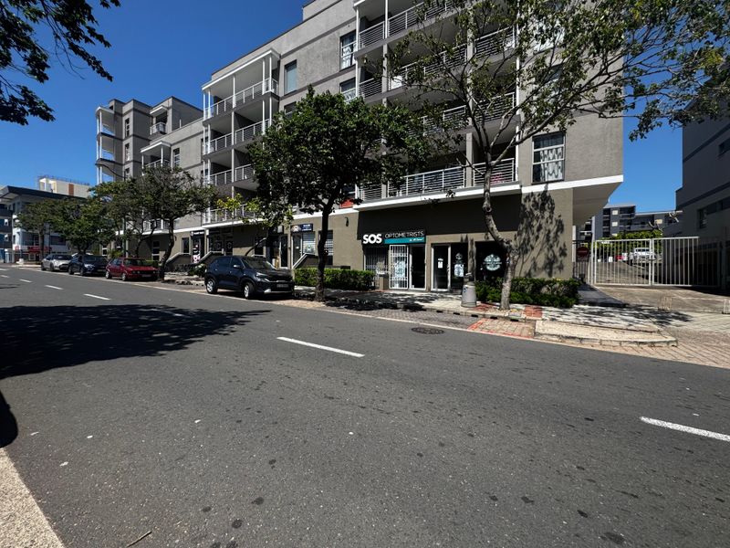 Street-level view of a modern multi-story retail and residential building featuring commercial storefronts with security gates and balcony access.