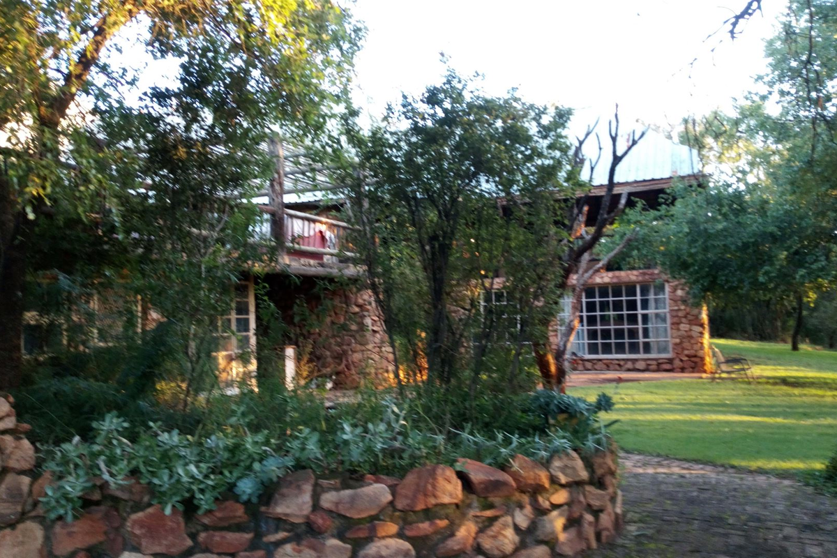 Rear view of a rustic stone-clad farmhouse featuring a timber balcony, grid-pattern windows, and a landscaped garden with a stone retaining wall.
