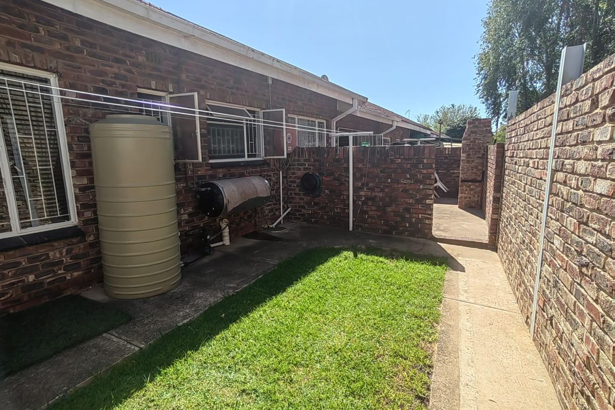 Rear courtyard area featuring a face brick wall, built-in braai, paved walkway, small lawn, and external water tank and geyser installation.