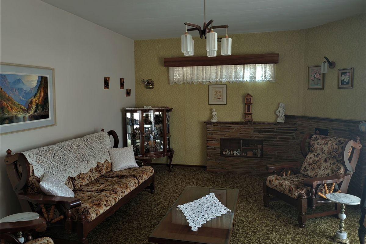 A dated living room featuring a stone fireplace, patterned wallpaper, and carpet flooring, offering potential for a modern renovation.