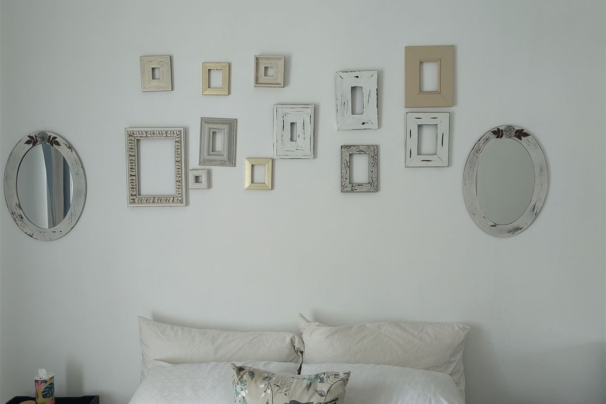 A bedroom interior featuring a wall decorated with a collection of various empty picture frames and two oval mirrors.