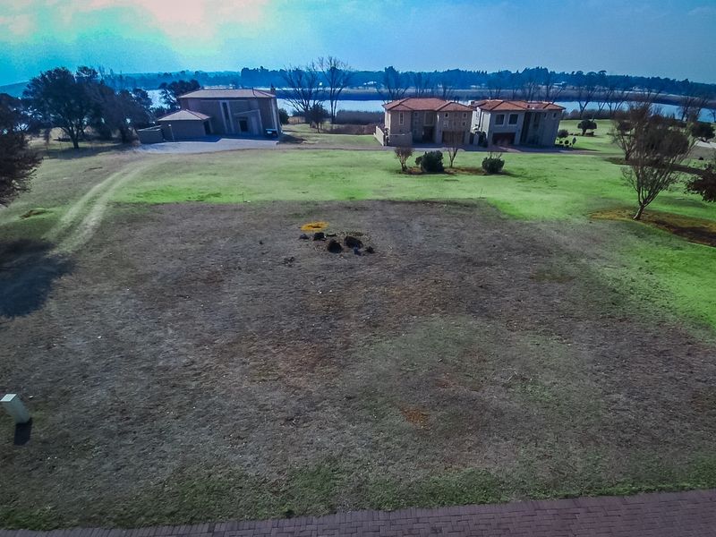 An aerial view of a vacant residential plot featuring open land with surrounding neighborhood properties and a scenic water view in the distance.
