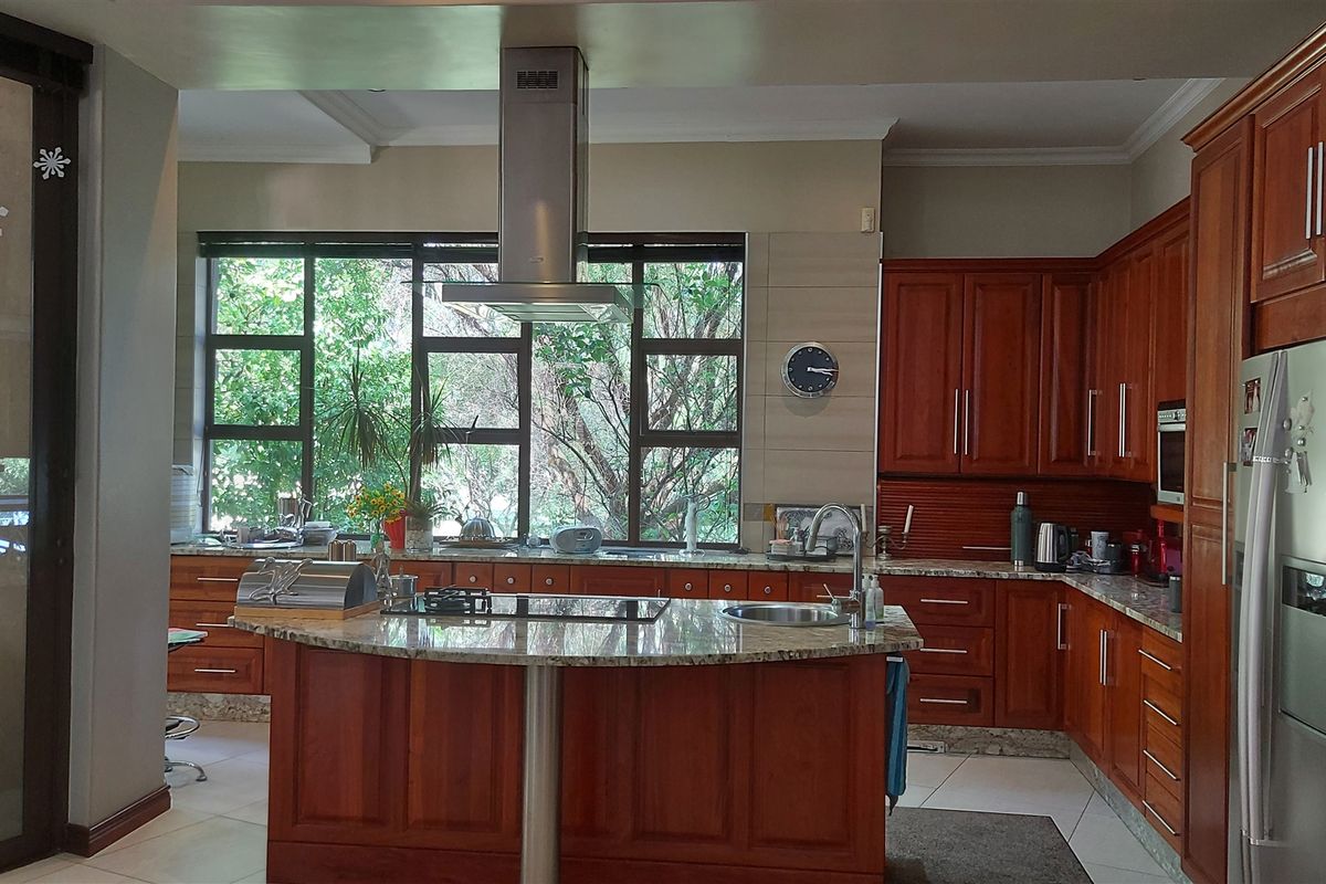 A spacious kitchen featuring warm-toned wooden cabinetry, granite countertops, a central island with a built-in hob, and a stainless steel extractor fan.