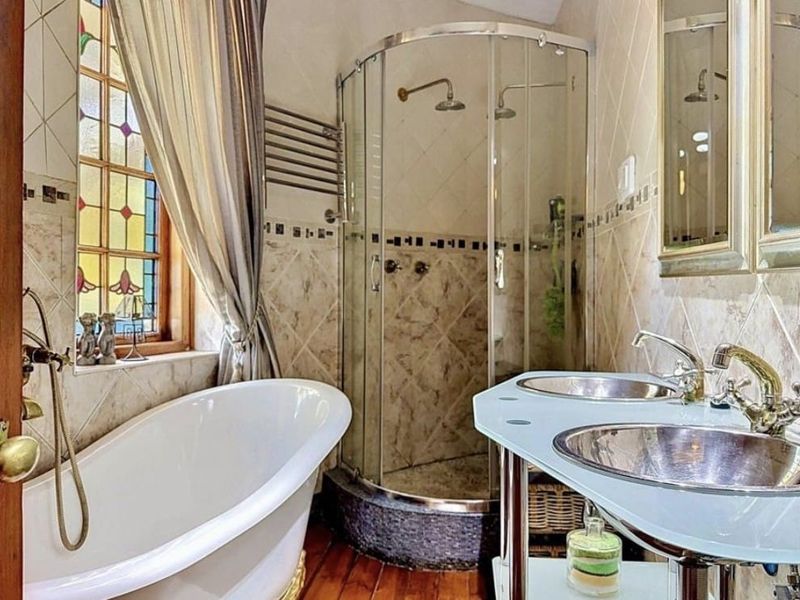 A well-appointed bathroom featuring a freestanding bathtub, a corner shower enclosure, and a double vanity with metallic sinks, complemented by a decorative stained glass window.