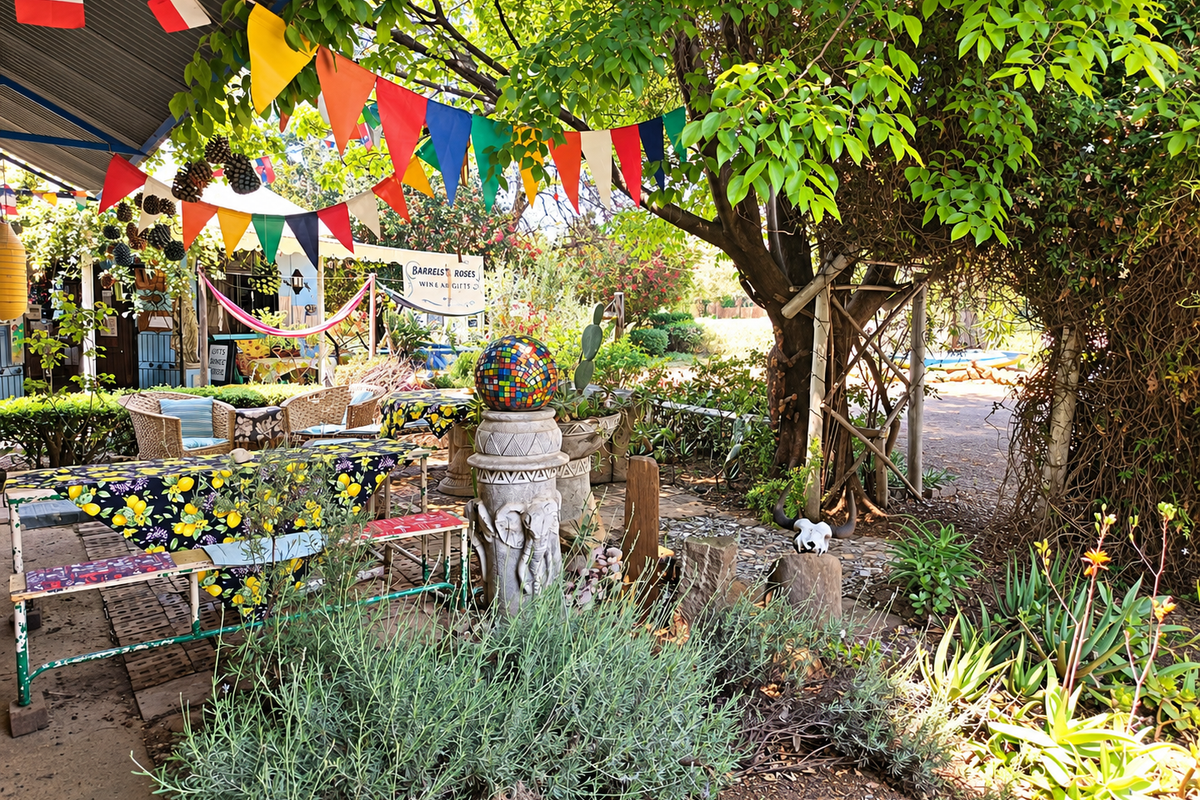 A shaded outdoor patio area featuring a corrugated metal roof, paved flooring, and established garden surroundings, suitable for commercial outdoor seating.