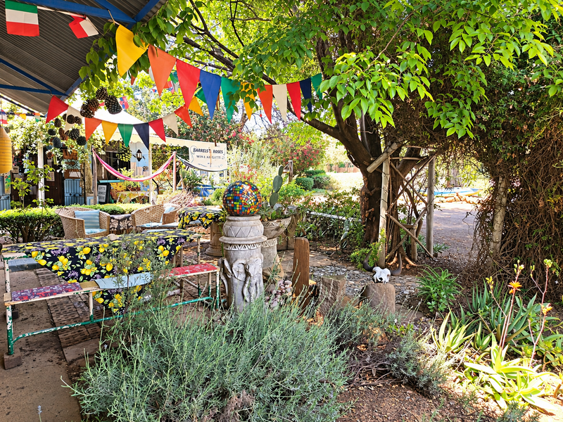 A shaded outdoor patio area featuring a corrugated metal roof, paved flooring, and established garden surroundings, suitable for commercial outdoor seating.