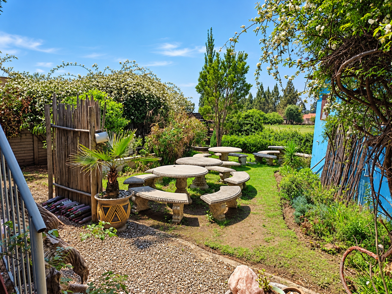A landscaped garden area featuring stone tables and benches, surrounded by mature greenery, bamboo fencing, and a gravel pathway.