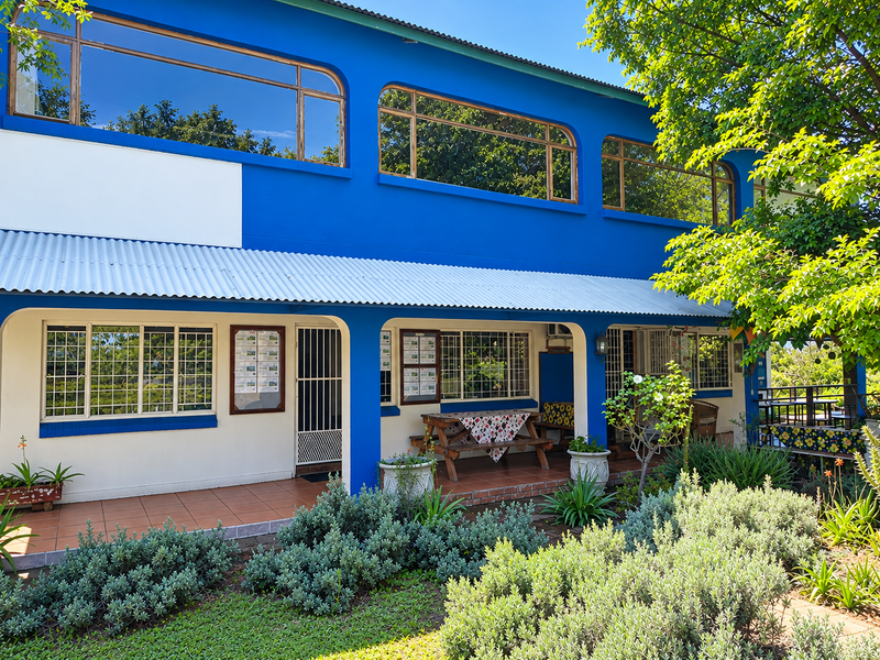A two-story commercial building exterior featuring a blue facade, large windows, a covered tiled patio area, and a well-maintained garden.