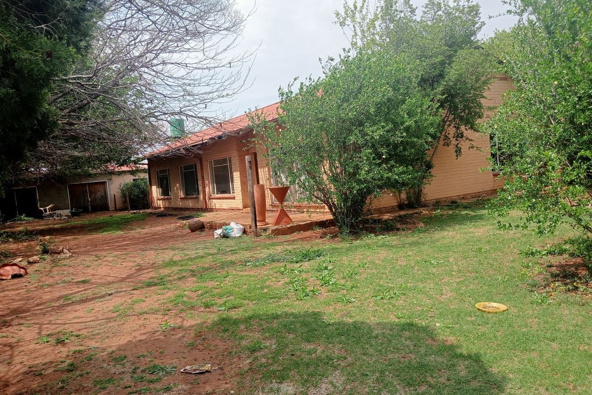 Rear view of a farm-style residence featuring a brick facade, corrugated metal roof, and an attached concrete patio area set within a large garden.