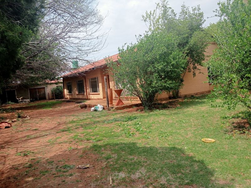 Rear view of a farm-style residence featuring a brick facade, corrugated metal roof, and an attached concrete patio area set within a large garden.