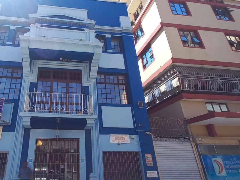 Street-level view of a multi-story commercial office building featuring a blue facade, balcony, and ground-floor security gates.