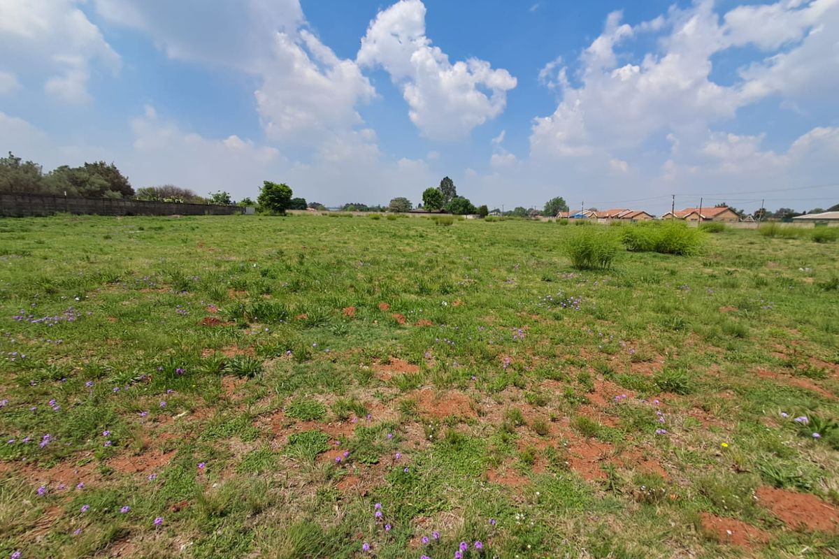 A wide-angle view of a vacant commercial land plot featuring natural grass coverage and a perimeter boundary wall in the background.