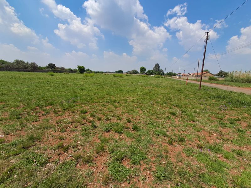 A wide-angle view of a vacant commercial land plot featuring grassy terrain, boundary walling, and existing utility infrastructure along the roadside.