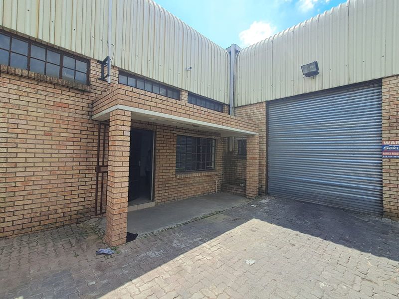 Industrial property exterior featuring a brick facade, a large roller shutter door, and a covered entrance area with paved driveway access.