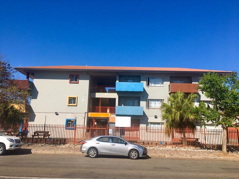 Front elevation of a multi-story apartment building featuring balconies, metal perimeter fencing, and a tiled roof, viewed from the street.
