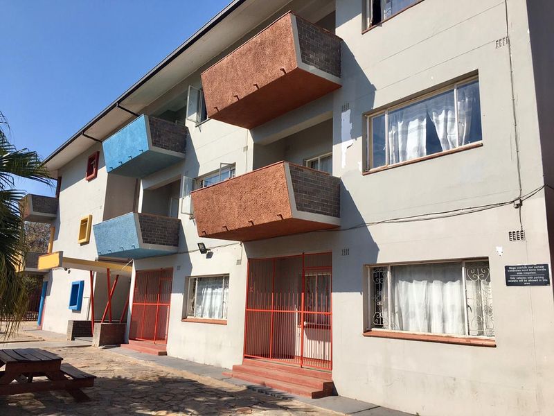 Exterior view of a multi-story apartment building featuring balconies, security gates, and paved ground area.