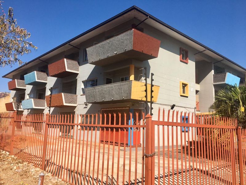 Exterior view of a multi-story apartment building featuring private balconies, security fencing, and a gated entrance.