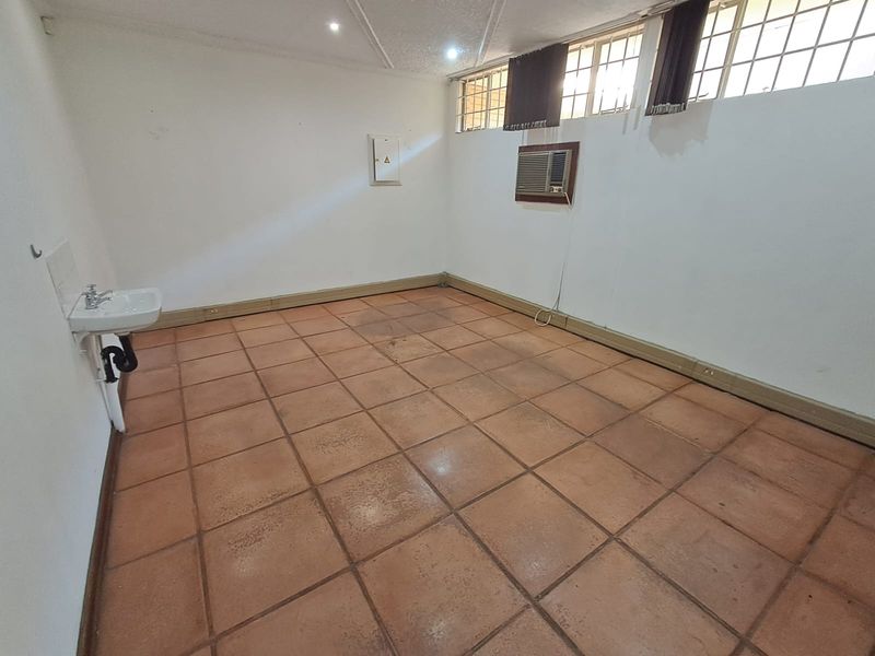 An empty office space featuring tiled flooring, a wall-mounted basin, and a window-mounted air conditioning unit.