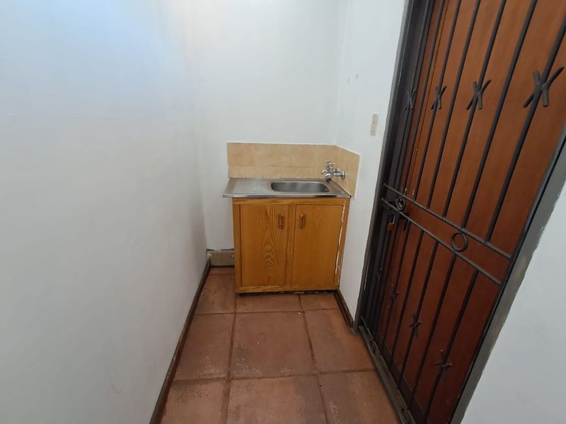 A compact utility or scullery area featuring a stainless steel sink with a wooden base cabinet, tiled splashback, and a secure metal entrance gate.