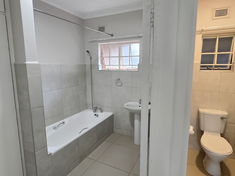 A functional bathroom featuring a pedestal basin, built-in bathtub with shower mixer, and a separate toilet area with neutral wall and floor tiling.