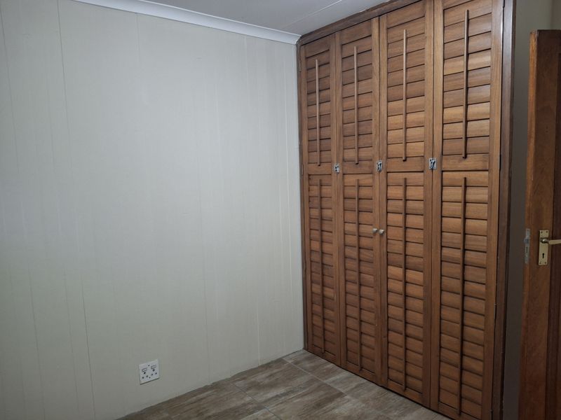A clean, empty bedroom featuring built-in wooden louvered wardrobes, tiled flooring, and neutral wall finishes.