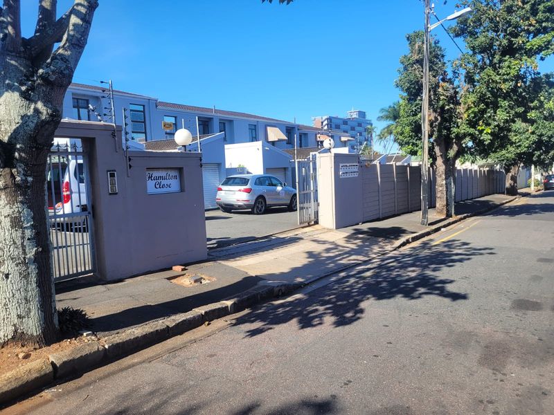 Street-level view of the Hamilton Close townhouse complex entrance, featuring secure perimeter walling, electric fencing, and a gated driveway access point.