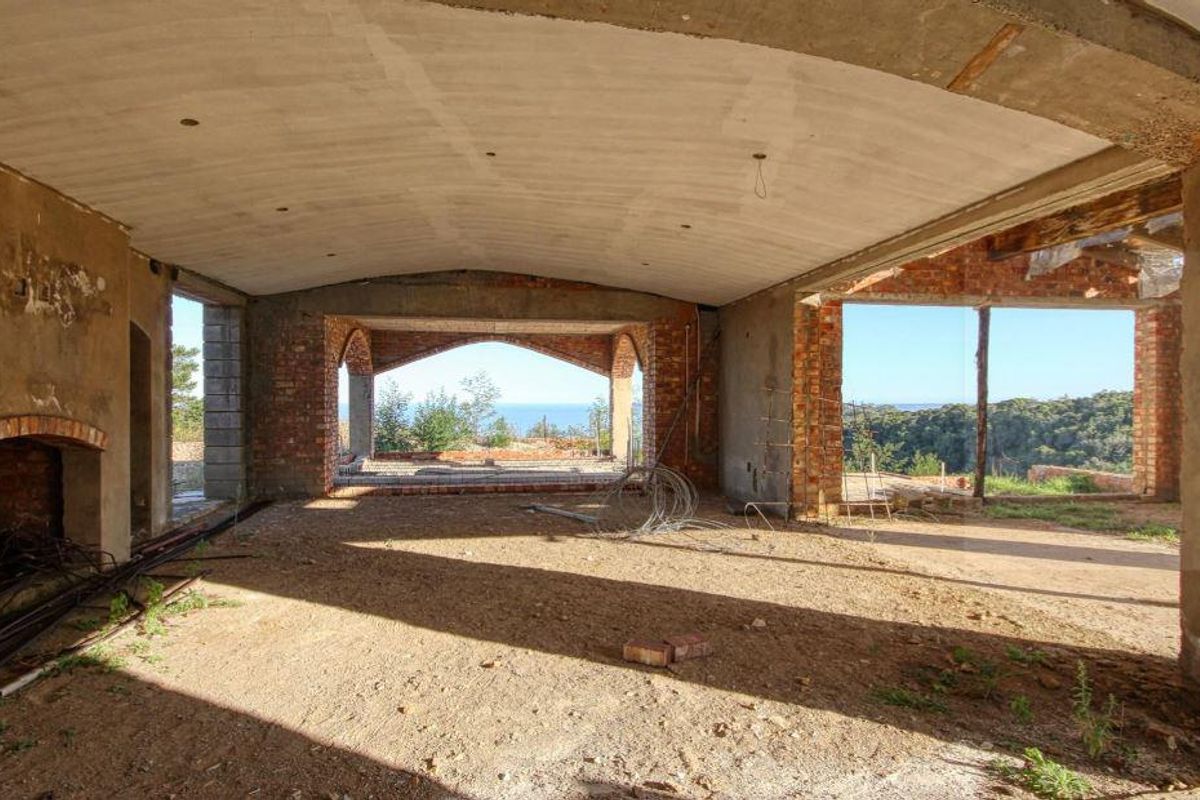 An unfinished living area in shell condition featuring a brick fireplace, vaulted ceiling, and large openings with scenic views, awaiting completion and interior finishes.