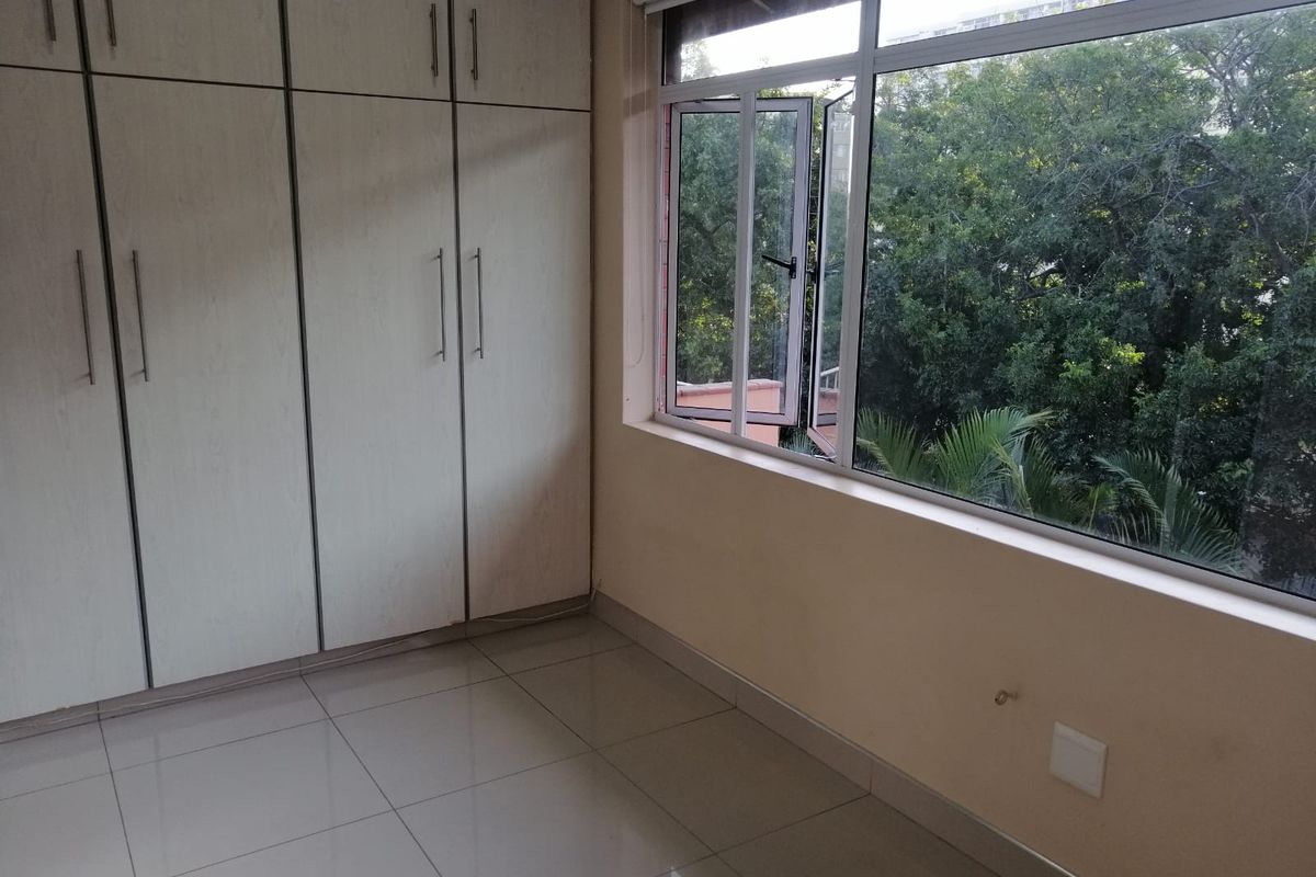 A bright, empty bedroom featuring tiled flooring, ample built-in cupboard storage, and large windows overlooking lush greenery.