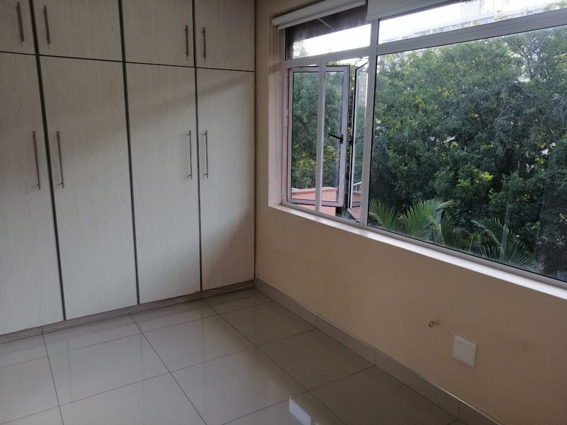 A bright, empty bedroom featuring tiled flooring, ample built-in cupboard storage, and large windows overlooking lush greenery.