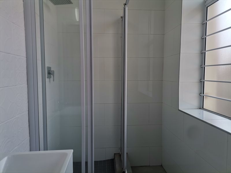 A clean, modern bathroom featuring a glass shower enclosure, tiled walls, and a window with security bars.