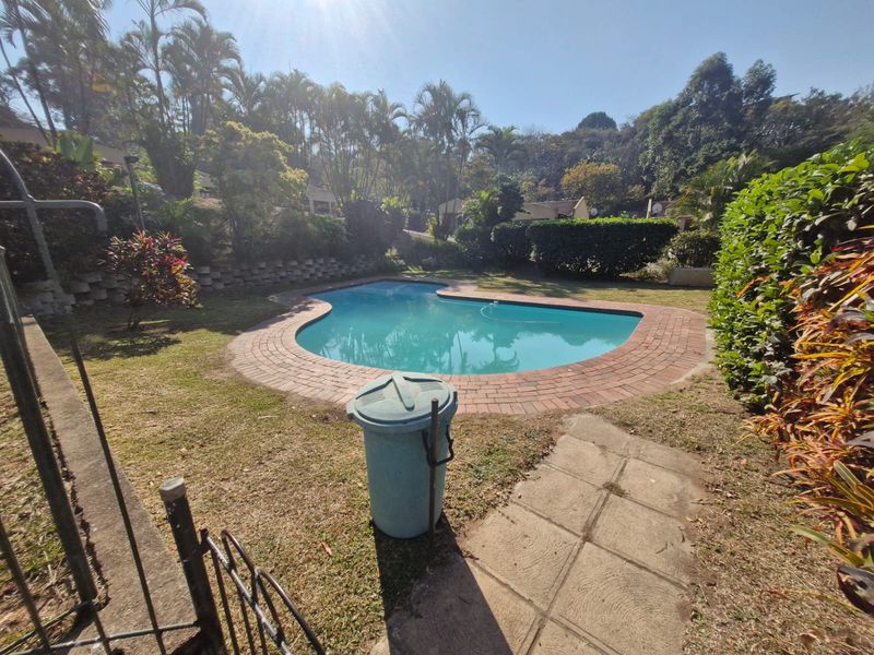 A private swimming pool area featuring brick coping, a paved walkway, and established garden landscaping in a townhouse complex setting.