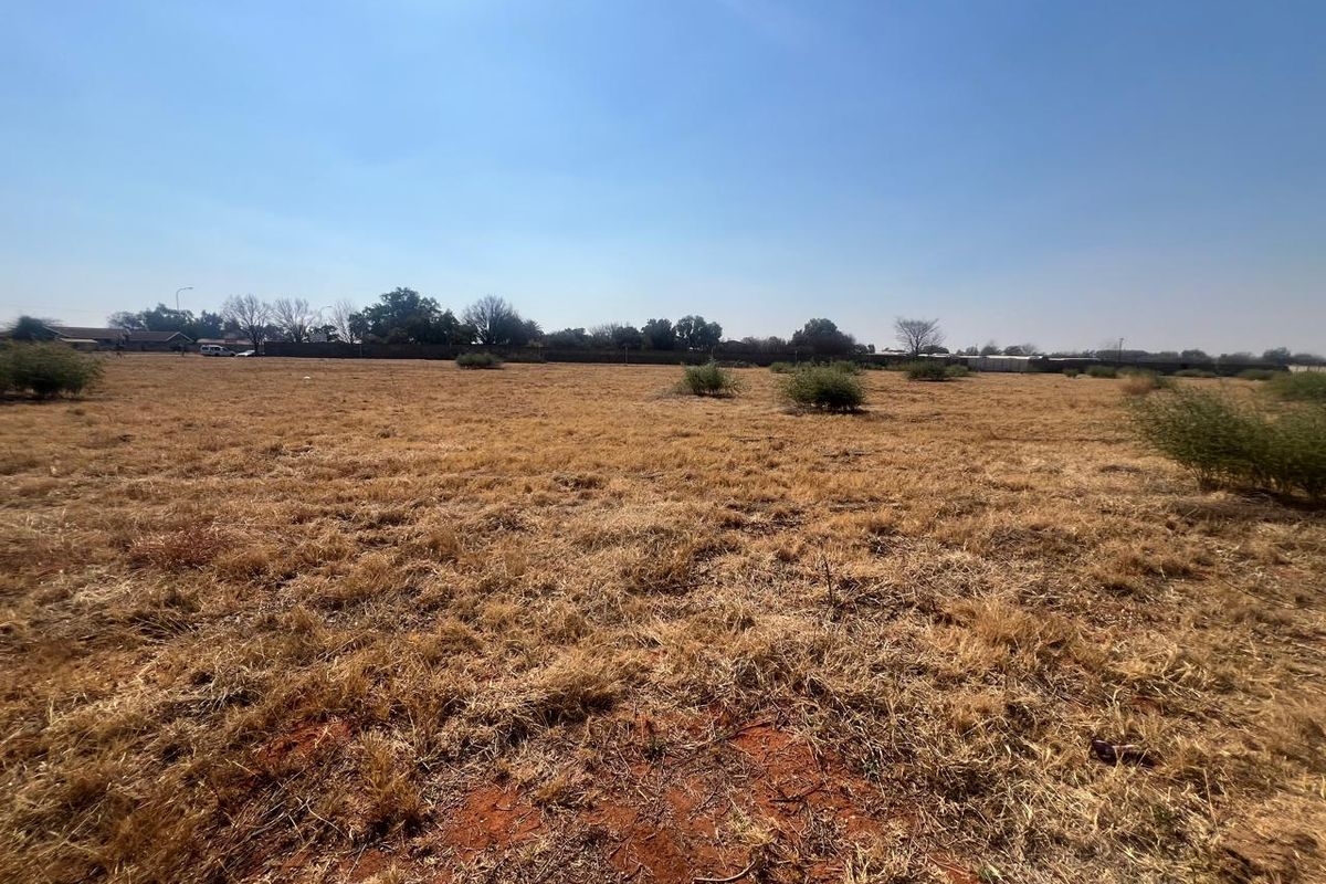 A wide-angle view of a vacant residential plot featuring dry grass and a perimeter boundary wall in the background.
