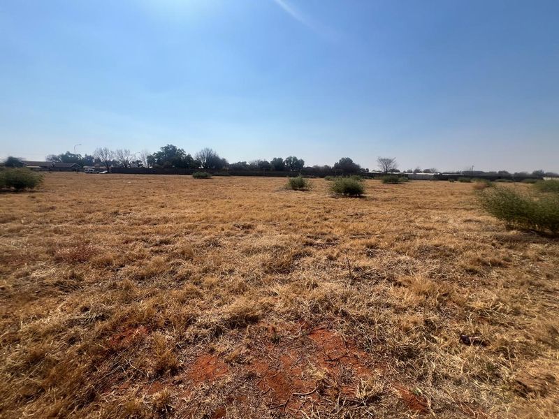 A wide-angle view of a vacant residential plot featuring dry grass and a perimeter boundary wall in the background.