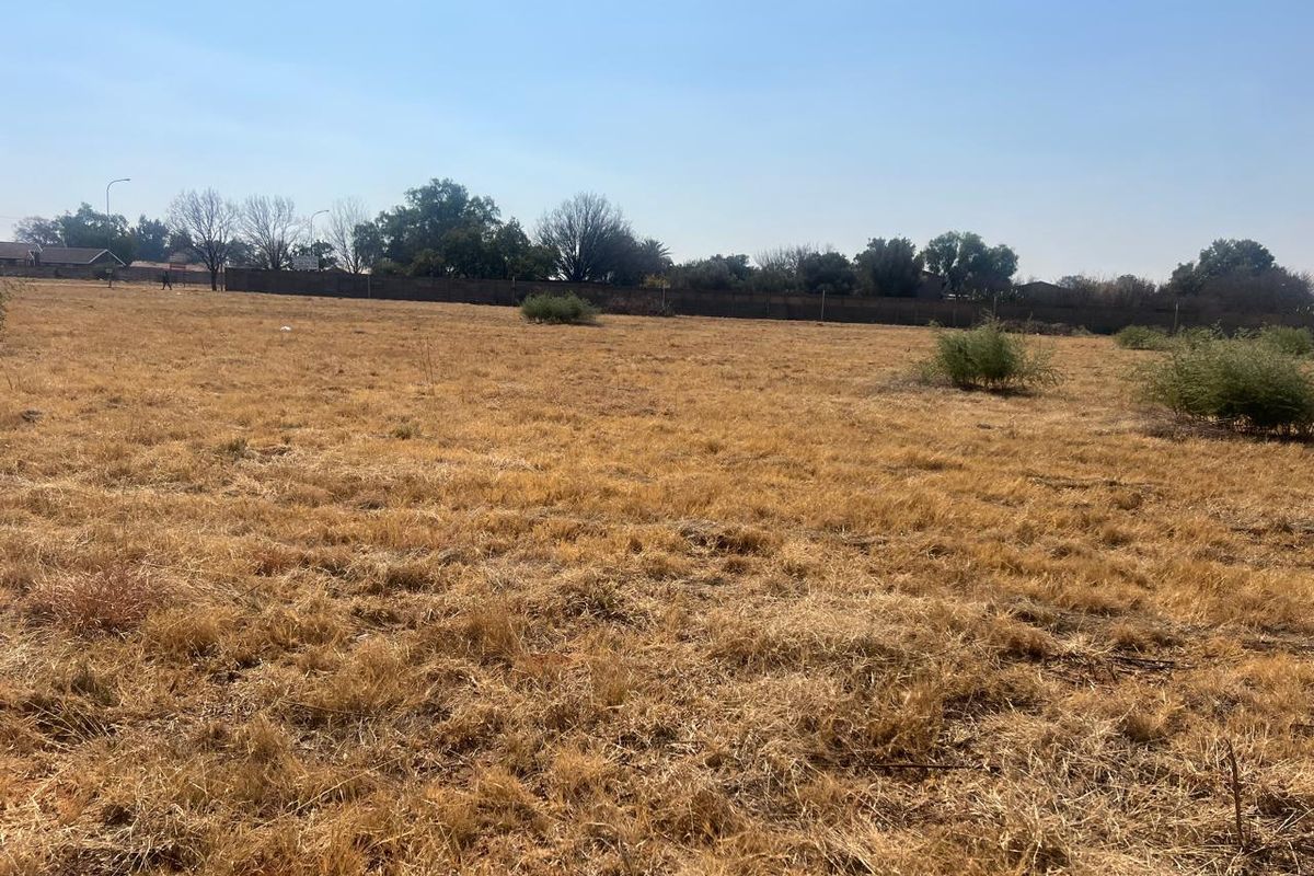 A wide, level vacant residential plot featuring dry grass and a perimeter boundary wall, offering a blank canvas for future development.