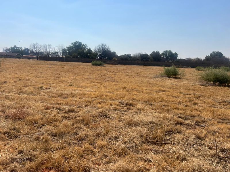 A wide, level vacant residential plot featuring dry grass and a perimeter boundary wall, offering a blank canvas for future development.