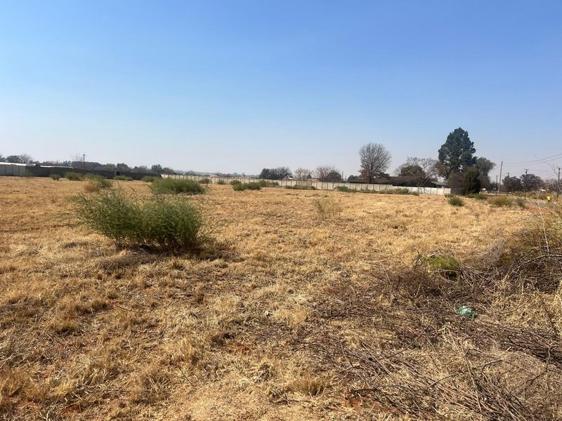 A wide-angle view of a vacant residential plot featuring dry grass and a perimeter boundary wall in the background.