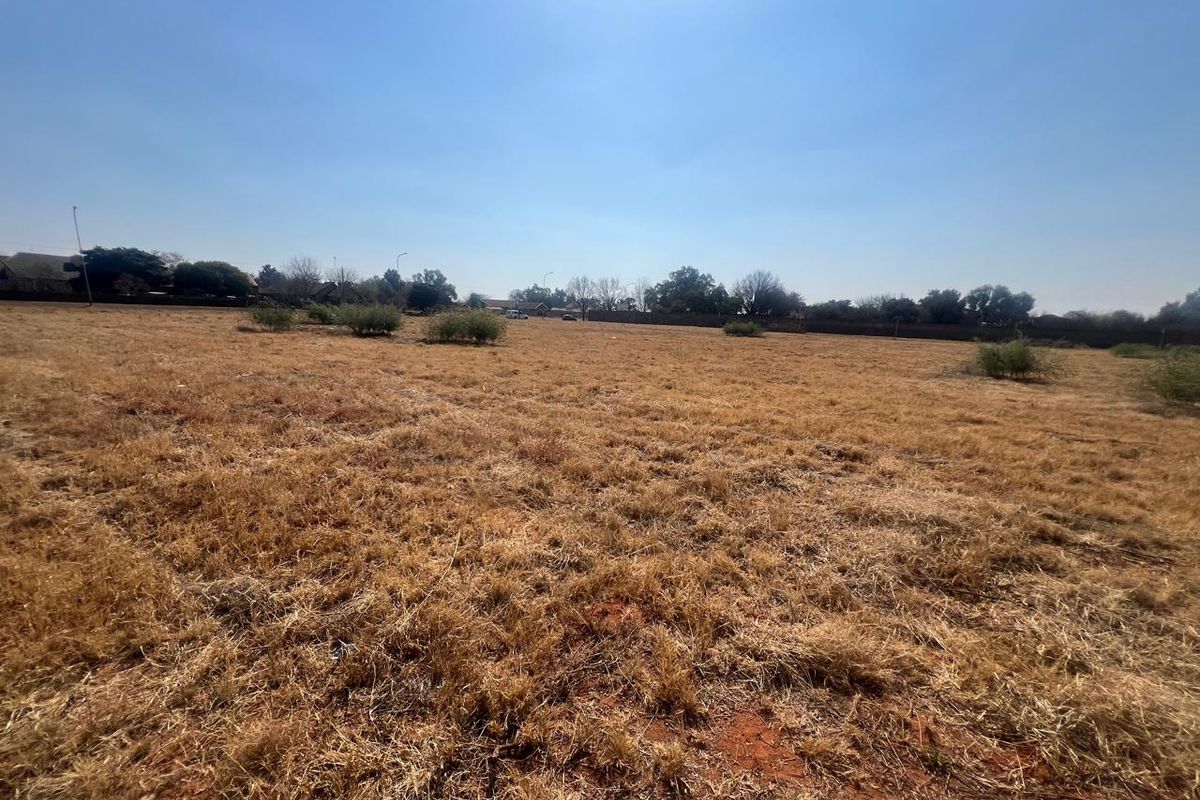 A wide-angle view of a vacant residential plot featuring dry grass and a perimeter wall in the background.