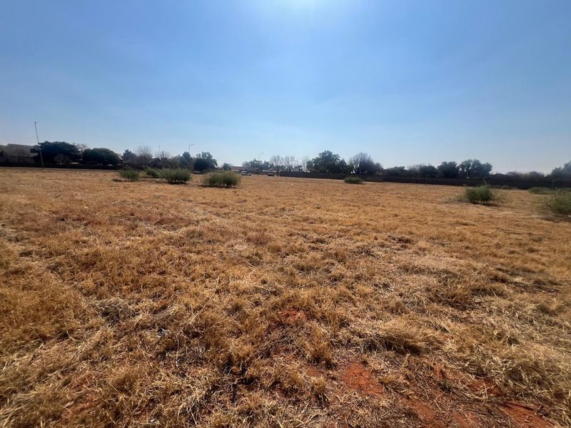 A wide-angle view of a vacant residential plot featuring dry grass and a perimeter wall in the background.