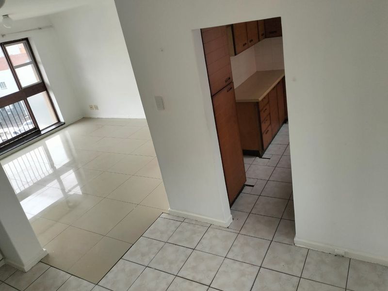 An open-plan living area featuring tiled floors and a separate kitchen space with built-in cabinetry, offering a functional layout in a bright, vacant apartment.