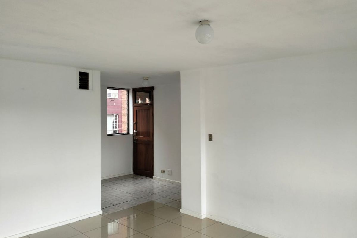An empty, neutrally painted living area featuring tiled flooring, a ceiling light fixture, and a wooden door leading to an adjacent space.