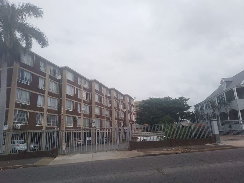 Street view of a multi-story residential flat complex featuring brick facade, secure gated entrance, and perimeter fencing.