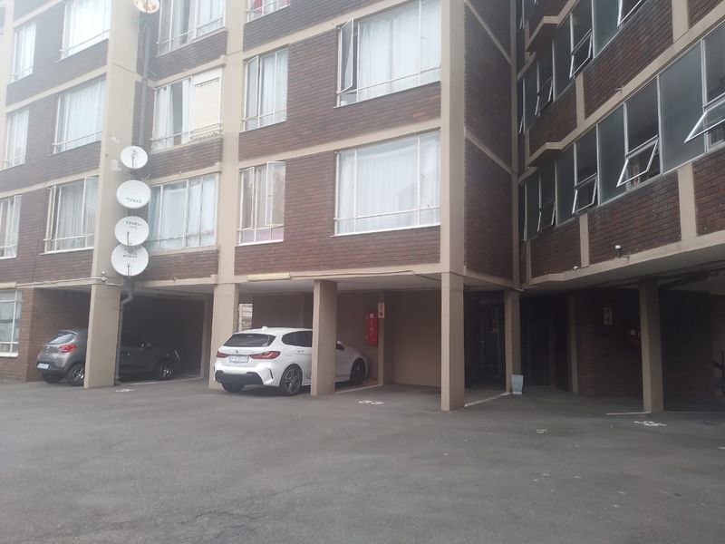Exterior view of a multi-story apartment building featuring a brick facade, concrete support pillars, and a paved ground-level parking area.