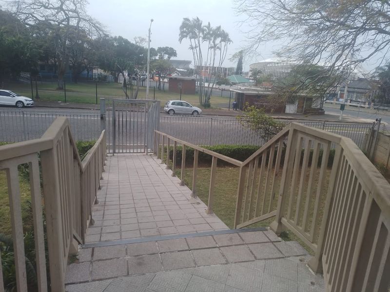 A paved pedestrian walkway with metal security railings leading to a gated entrance, overlooking a street and surrounding neighbourhood area.