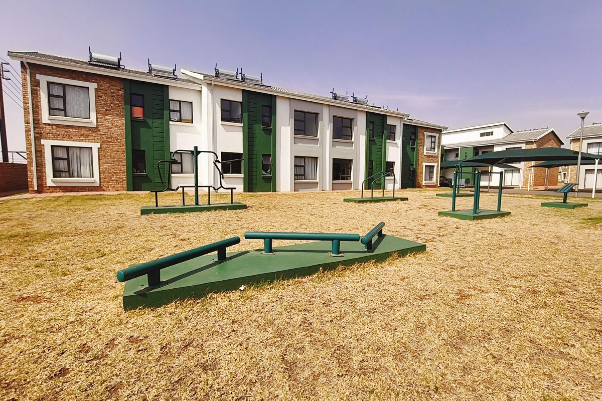 Outdoor communal area featuring fixed exercise equipment and landscaped grounds set against a multi-story apartment complex with solar water heating systems.