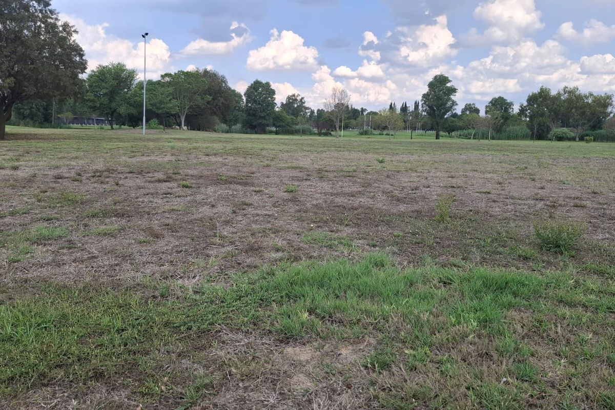 A wide, open residential plot of land featuring natural grass coverage and mature trees in the background.