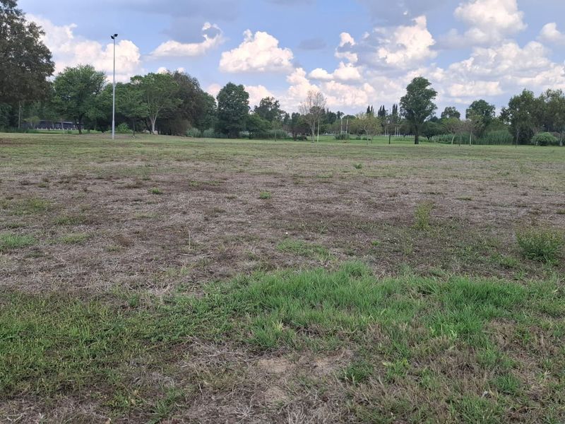A wide, open residential plot of land featuring natural grass coverage and mature trees in the background.