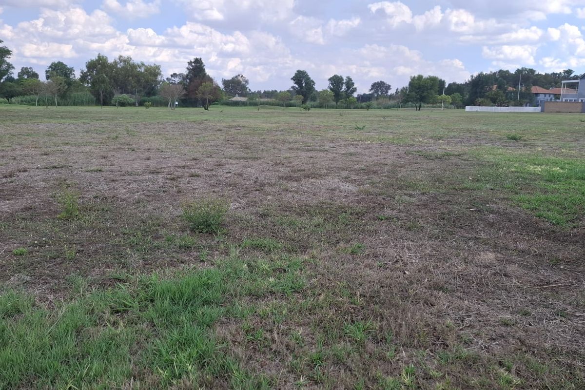 A wide, level vacant residential plot featuring open grassland with surrounding mature trees and nearby residential development.